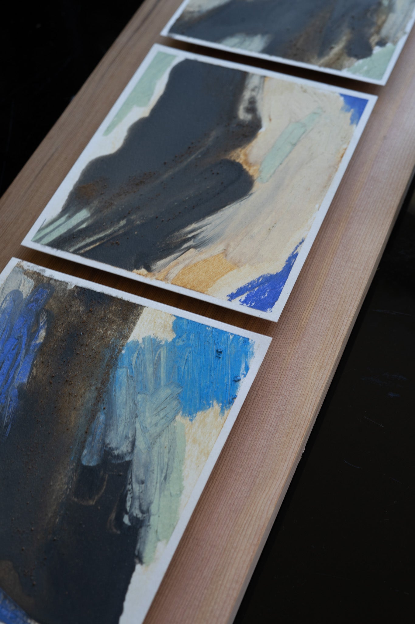 Threeabstract art pieces on a wooden panel with black, blue, and beige colors.
